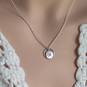 May include: A silver-toned necklace featuring a circular pendant with a compass rose design. The pendant consists of a small disc engraved with cardinal directions N, E, S, and W, suspended within a thin, plain silver-toned circular ring.