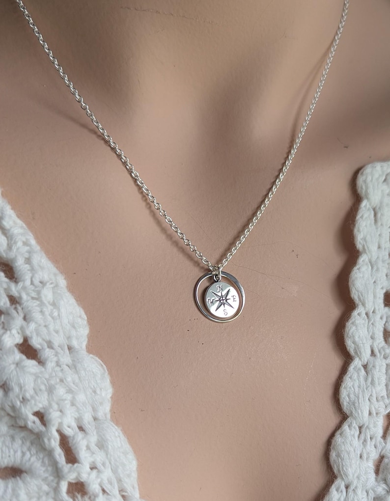 May include: A silver-toned necklace featuring a circular pendant with a compass rose design. The pendant consists of a small disc engraved with cardinal directions N, E, S, and W, suspended within a thin, plain silver-toned circular ring.