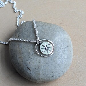 May include: A silver-toned necklace featuring a circular pendant with a compass rose design. The pendant is suspended from a delicate silver chain and includes a small, thin circular ring frame surrounding the compass charm.
