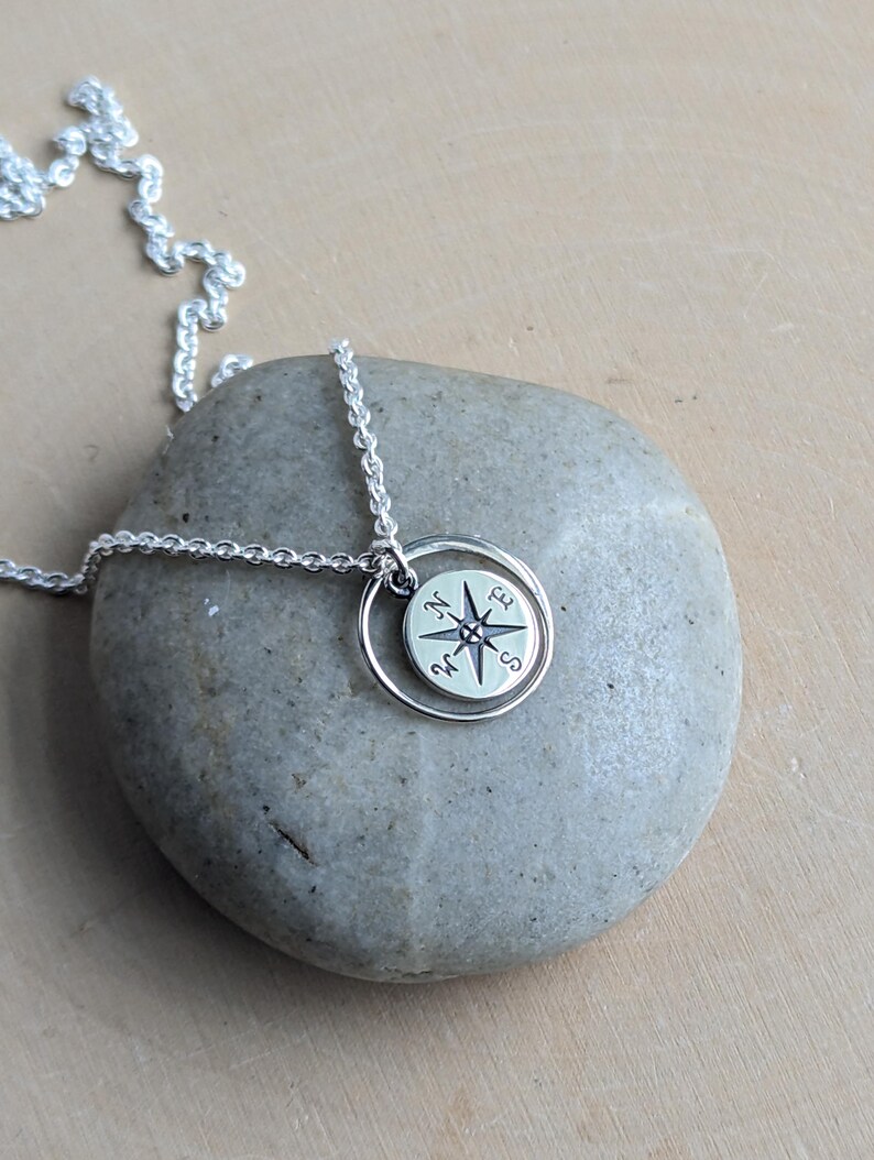 May include: A silver-toned necklace featuring a circular pendant with a compass rose design. The pendant is suspended from a delicate silver chain and includes a small, thin circular ring frame surrounding the compass charm.