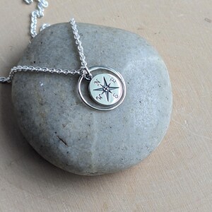 May include: A silver necklace featuring a circular pendant with an engraved compass rose design. The pendant is suspended from a delicate silver chain and displays the cardinal directions N, E, S, and W clearly marked on its polished surface.