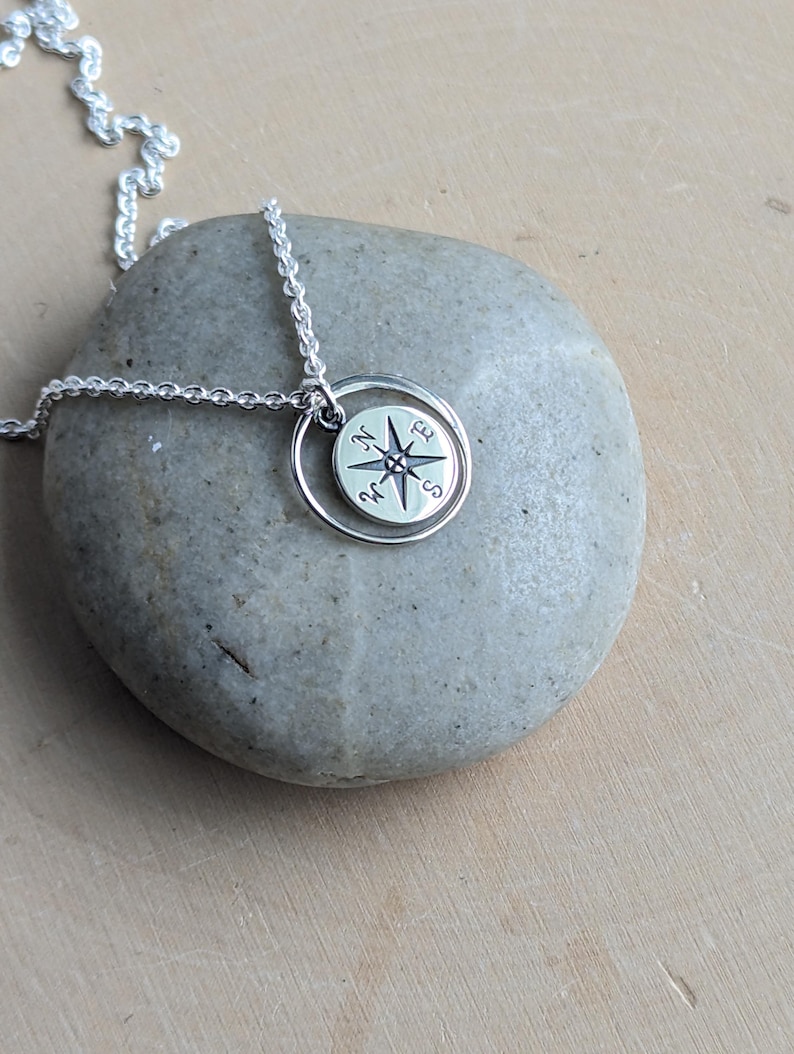 May include: A silver necklace featuring a circular pendant with an engraved compass rose design. The pendant is suspended from a delicate silver chain and displays the cardinal directions N, E, S, and W clearly marked on its polished surface.