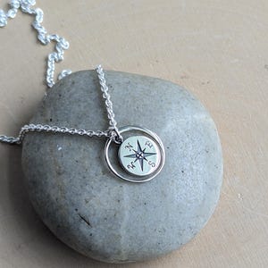 May include: A silver necklace featuring a circular pendant with a compass rose design. The pendant consists of a small disc engraved with cardinal directions N, E, S, and W, suspended within a thin, plain silver ring on a delicate chain.