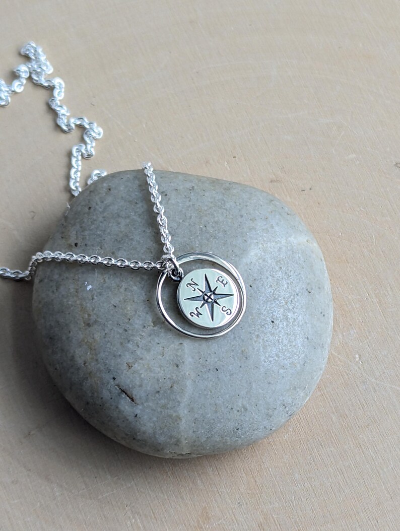 May include: A silver necklace featuring a circular pendant with a compass rose design. The pendant consists of a small disc engraved with cardinal directions N, E, S, and W, suspended within a thin, plain silver ring on a delicate chain.
