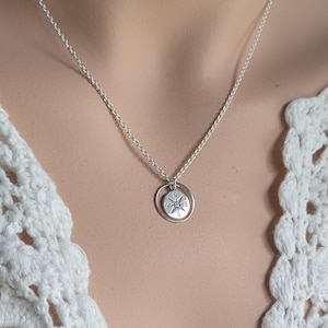 May include: A silver-toned necklace featuring a delicate chain and a circular pendant. The pendant consists of a small, flat disc engraved with a compass rose design, suspended within a simple, thin silver-coloured metal ring frame.