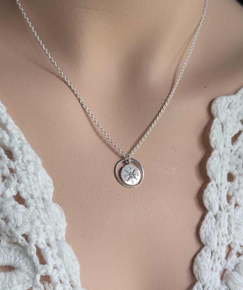 May include: A silver-toned necklace featuring a delicate chain and a circular pendant. The pendant consists of a small, flat disc engraved with a compass rose design, suspended within a simple, thin silver-coloured metal ring frame.