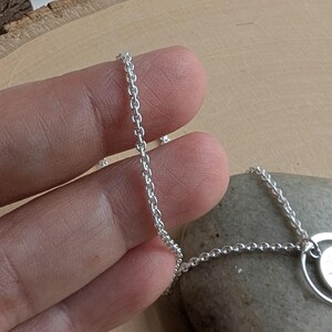 May include: A silver-coloured metal chain necklace featuring a delicate link design. The chain is connected to a circular pendant component, showcasing a polished, reflective surface that catches the light, creating a bright and metallic appearance.