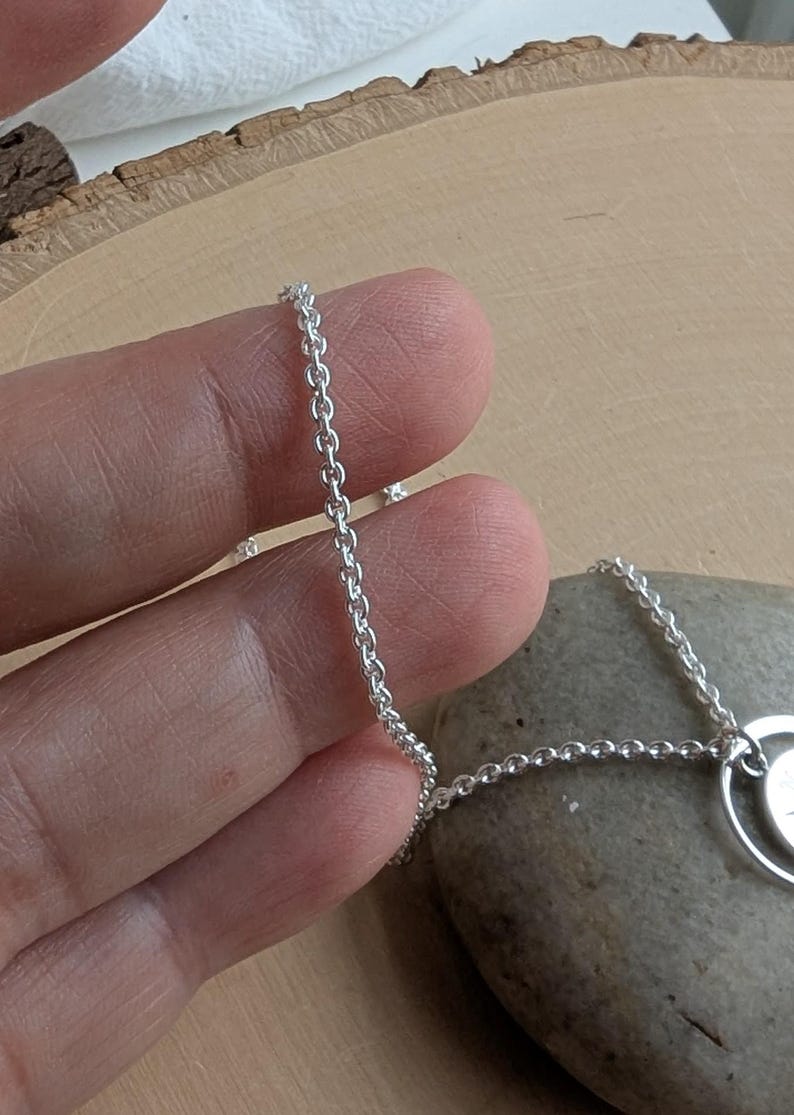 May include: A silver-coloured metal chain necklace featuring a delicate link design. The chain is connected to a circular pendant component, showcasing a polished, reflective surface that catches the light, creating a bright and metallic appearance.