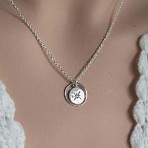 May include: A silver-toned necklace featuring a delicate chain and a circular pendant. The pendant consists of a thin outer ring surrounding a smaller, round charm engraved with a compass rose design, including the letters N, S, E, and W.