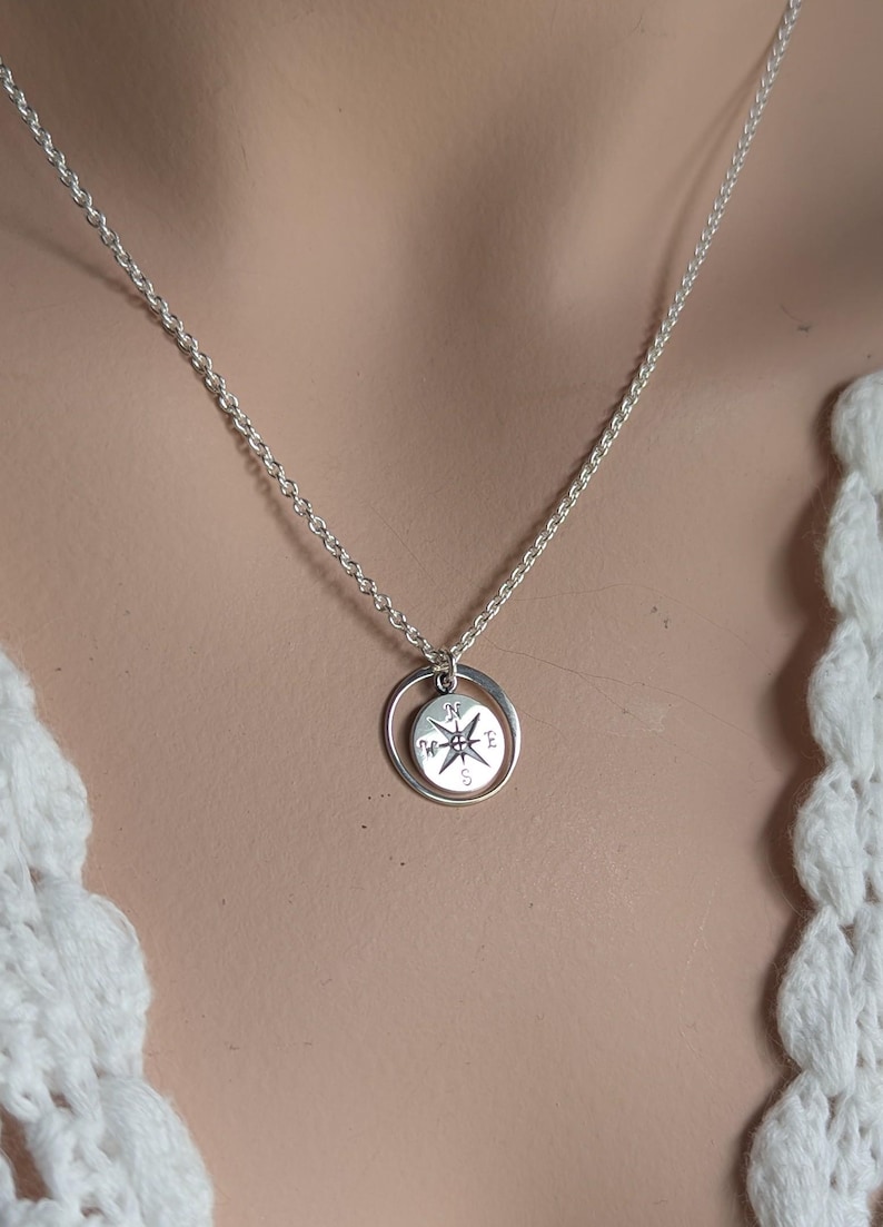 May include: A silver-toned necklace featuring a delicate chain and a circular pendant. The pendant consists of a thin outer ring surrounding a smaller, round charm engraved with a compass rose design, including the letters N, S, E, and W.