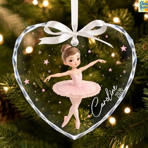 May include: A heart-shaped clear acrylic Christmas ornament featuring a cartoon ballerina in a pink tutu. The ornament is decorated with pink stars and snowflakes, includes a white ribbon for hanging, and displays the name Caroline and 2026.