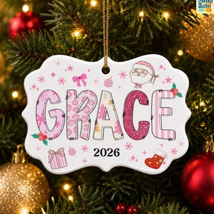 May include: A white, scalloped-edge ceramic ornament featuring the name GRACE in pink and white patterned letters. The design includes a Santa Claus face, a gift box, a stocking, and the year 2026 in black text, all accented with pink stars.