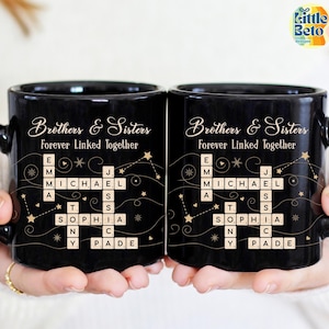 May include: Two black ceramic mugs with the text "Brothers & Sisters Forever Linked Together." Each mug features a crossword puzzle design with names. The mugs are held by a person.