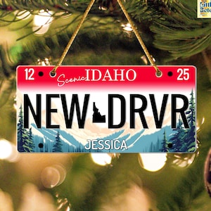 May include: A rectangular Christmas ornament resembling an Idaho license plate. The ornament features the text "Scenic IDAHO" and "NEW DRVR" with a mountain and forest scene. The name "JESSICA" is printed at the bottom.