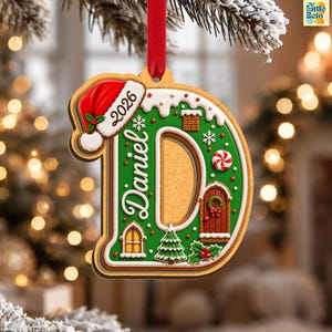 May include: A gingerbread-style letter D Christmas ornament featuring the name Daniel and the year 2026. The ornament is decorated with a red Santa hat, white icing, a brown door, a window, a green Christmas tree, and red holly berries.