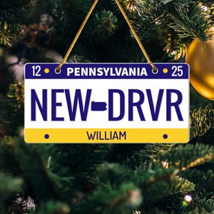 May include: A license plate ornament with the text "PENNSYLVANIA" and "NEW-DRVR" in blue on a white background. The name "WILLIAM" is printed in black. The ornament is hanging from a gold string.