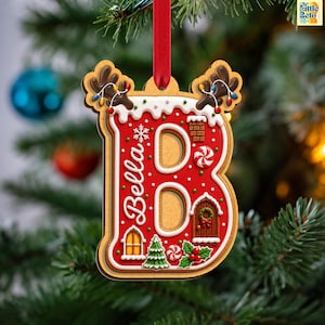 May include: A letter B shaped Christmas ornament with a gingerbread texture, red icing, and white trim. It features the name Bella, a small house, a chimney, a Christmas tree, holly, and reindeer antlers with colorful lights, hanging by a red ribbon.