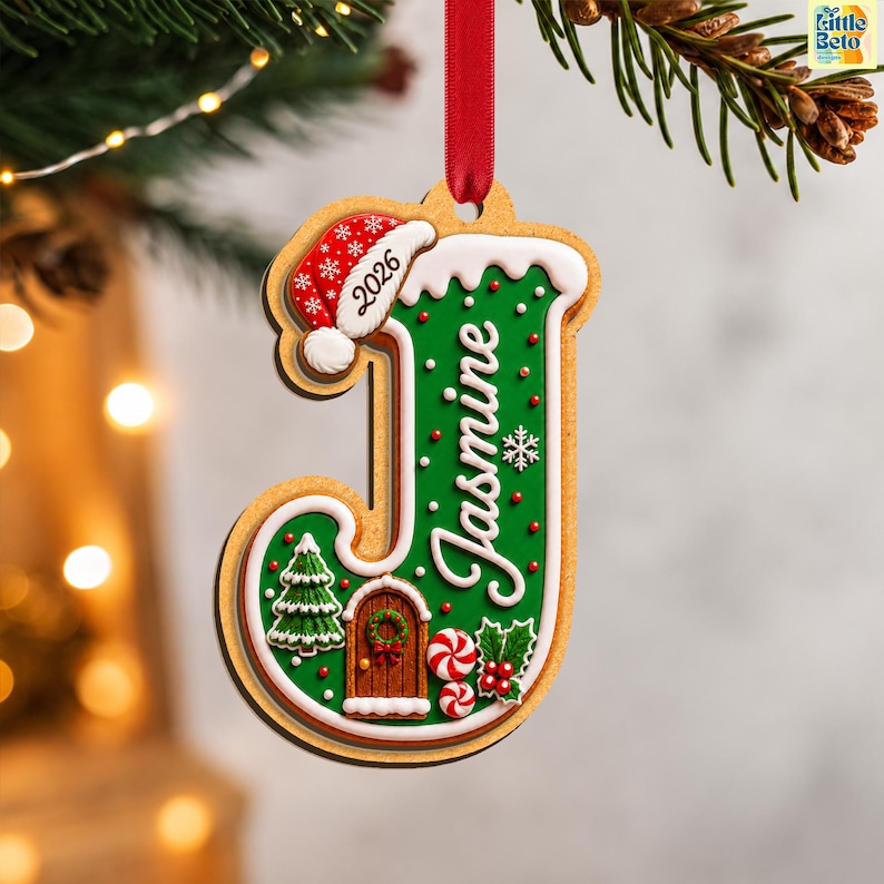 May include: A letter J-shaped Christmas ornament featuring a green gingerbread-style design with white icing trim. It includes a red Santa hat with the year 2026, the name Jasmine in white script, a small brown door, a tree, and peppermint candies.
