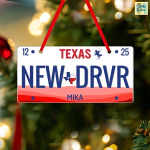 May include: A rectangular Christmas ornament resembling a Texas license plate. The ornament features the text "TEXAS" in red, "NEW DRVR" in blue, and "MIKA" in white. It is adorned with a red and white border and a red ribbon for hanging.