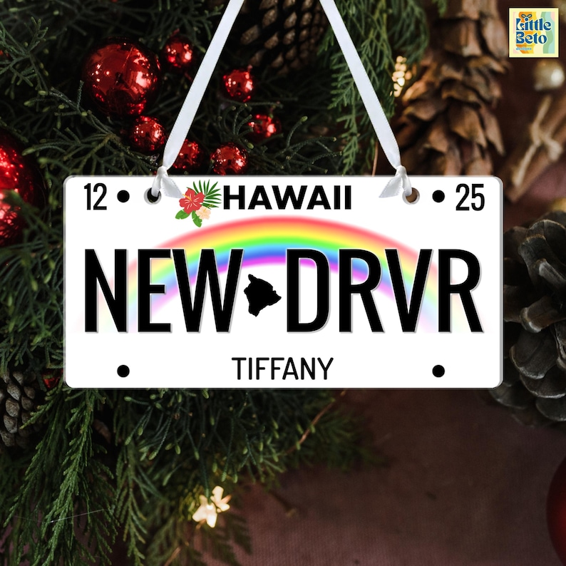 May include: A white Christmas ornament shaped like a license plate, with the text "NEW DRVR" and "HAWAII". It features a rainbow, a Hawaiian island silhouette, and the name "TIFFANY". The ornament is decorated with a white ribbon.