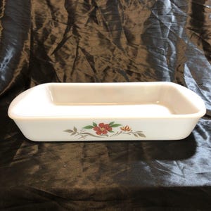 Puede incluir: Una fuente de horno rectangular de vidrio blanco con esquinas redondeadas. El lado exterior presenta un motivo floral decorativo que consiste en una flor roja con hojas verdes y una flor de acento naranja más pequeña, unidas por tallos grises.