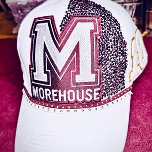 Könnte beinhalten: Weiße Baseballkappe mit einem großen, bordeauxroten und weißen Buchstaben "M" und dem Wort "MOREHOUSE" auf der Vorderseite. Die Kappe ist mit bordeauxfarbenen Pailletten und kleinen goldenen Nieten am Rand verziert.