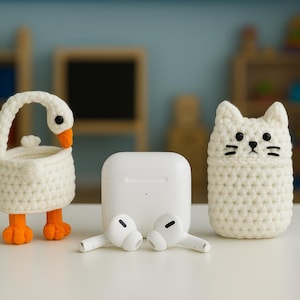 May include: A white crochet goose and cat case, along with a white charging case and earbuds. The goose has orange feet and beak, and the cat has black eyes and whiskers. The items are on a white surface.