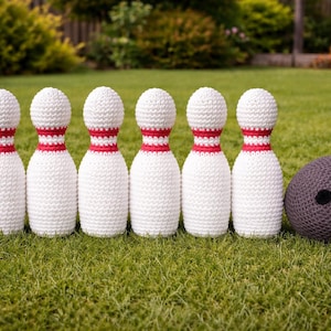 Peut inclure: Un ensemble de six quilles de bowling blanches crochetées avec des rayures rouges et une boule de bowling grise crochetée avec trois trous noirs, le tout disposé sur une surface herbeuse verte. Les quilles sont alignées, et la boule est sur le côté.