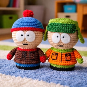 Pode incluir: Duas bonecas de crochê, uma usando um chapéu azul com um pompom vermelho e uma jaqueta marrom, a outra com um chapéu verde e uma jaqueta laranja. Elas estão em um tapete listrado azul e branco.