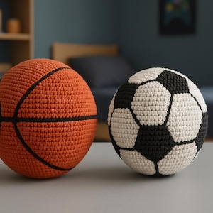 May include: Two crocheted sports balls: an orange basketball with black lines and a soccer ball with black and white pentagons. The balls are handmade and sit on a gray surface, suggesting a playful theme.