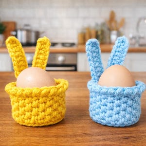 May include: Two handmade crochet egg holders, one yellow and one blue, each shaped like a basket with bunny ears. Each holder contains a brown egg. The items are on a wooden surface.