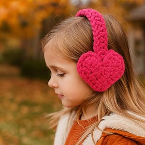 May include: A young person wearing bright pink, heart-shaped ear muffs. The ear warmers are made of a chunky knit material. The background is blurred, with fall foliage and a house visible.