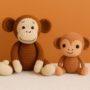 Puede incluir: Dos monos de ganchillo hechos a mano en tonos marrón y crema. El mono más grande tiene la cara, las manos y los pies de color crema claro. El mono más pequeño es de color marrón liso. Ambos tienen ojos negros y caras sonrientes.