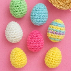 Op de afbeelding: Zeven gehaakte paaseieren in verschillende kleuren: groen, blauw, wit, roze en geel. Eén ei heeft horizontale strepen. De eieren liggen op een roze ondergrond, met een geweven nest op de achtergrond.