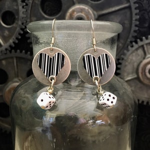 Può includere: Orecchini color argento con un design a cuore a forma di codice a barre in bianco e nero. Ogni orecchino presenta un piccolo dado bianco con punti neri pendenti. Gli orecchini sono appesi a ganci per orecchie color argento.