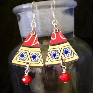 May include: A pair of triangle-shaped earrings made from recycled materials. The earrings feature a red, yellow and blue geometric pattern with a red bead dangling from each earring.