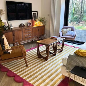 Op de afbeelding: Een woonkamer met een mid-century modern design. Een gestreept tapijt met goud-witte strepen en een geschulpte fuchsia rand. Een houten salontafel, een bruine fauteuil en een houten kast met een tv maken de kamer compleet.