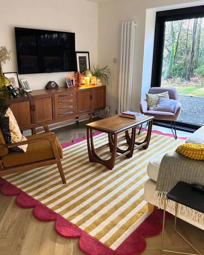 Op de afbeelding: Een woonkamer met een mid-century modern design. Een gestreept tapijt met goud-witte strepen en een geschulpte fuchsia rand. Een houten salontafel, een bruine fauteuil en een houten kast met een tv maken de kamer compleet.