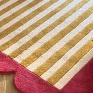 Op de afbeelding: Een close-up van een tapijt met een gestreept patroon in goud en wit. Het tapijt heeft een geschulpte, felroze rand. Het tapijt ligt op een houten vloer.