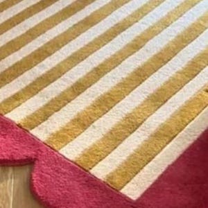 Op de afbeelding: Een close-up van een tapijt met een gestreept patroon in goud en wit. Het tapijt heeft een geschulpte, felroze rand. Het tapijt ligt op een houten vloer.