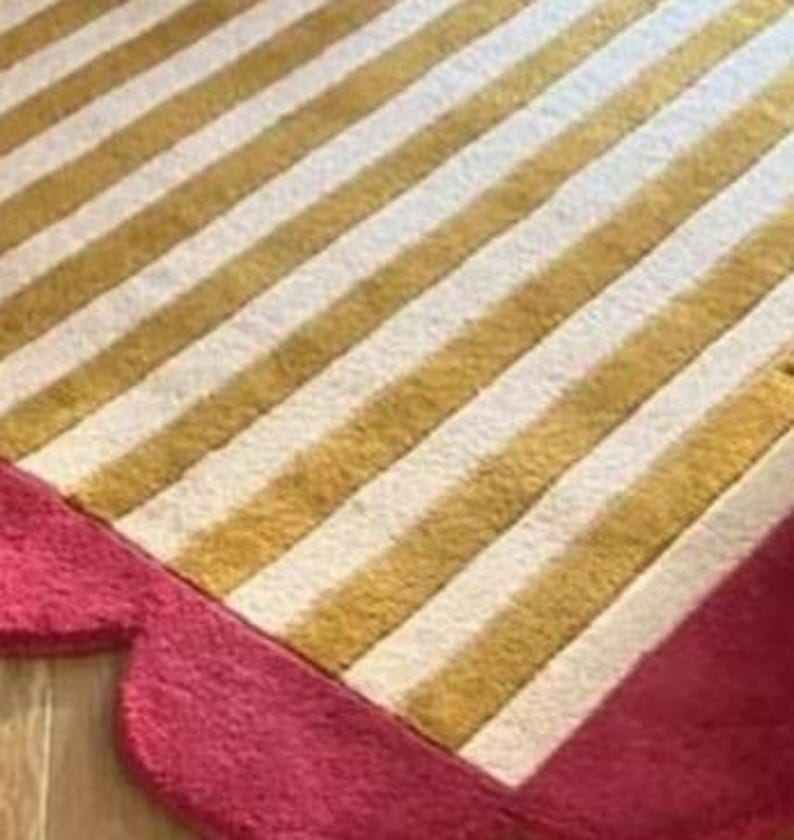 Op de afbeelding: Een close-up van een tapijt met een gestreept patroon in goud en wit. Het tapijt heeft een geschulpte, felroze rand. Het tapijt ligt op een houten vloer.