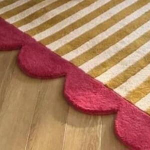 Op de afbeelding: Een tapijt met gouden en witte strepen, afgezet met een geschulpte rand in een dieprode kleur. Het tapijt ligt op een houten vloer, wat een contrast in texturen en kleuren cre&euml;ert.
