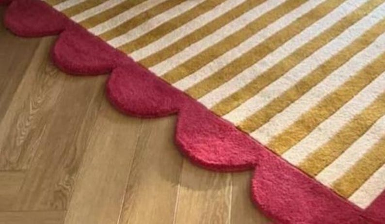 Op de afbeelding: Een tapijt met gouden en witte strepen, afgezet met een geschulpte rand in een dieprode kleur. Het tapijt ligt op een houten vloer, wat een contrast in texturen en kleuren cre&euml;ert.