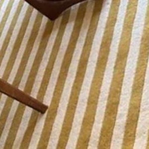 Op de afbeelding: Een close-up van een gestreept tapijt met afwisselende gouden en witte verticale strepen. Een deel van een houten stoelpoten is zichtbaar in de linkerbenedenhoek. Het tapijt heeft een getextureerd uiterlijk en een roze geschulpte rand.