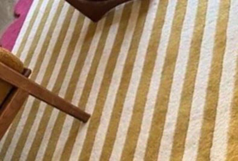 Op de afbeelding: Een close-up van een gestreept tapijt met afwisselende gouden en witte verticale strepen. Een deel van een houten stoelpoten is zichtbaar in de linkerbenedenhoek. Het tapijt heeft een getextureerd uiterlijk en een roze geschulpte rand.