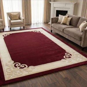 May include: A rectangular burgundy rug with a cream border and decorative scrollwork at the corners. The rug is on a wooden floor in a living room, with a sofa and chairs in the background. The rug's dimensions are not visible.