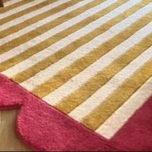 Op de afbeelding: Een gestreept tapijt met afwisselende gouden en witte strepen. Het tapijt heeft een geschulpte, roze rand. Het tapijt ligt op een houten vloer en een deel van een stoelpot is zichtbaar.