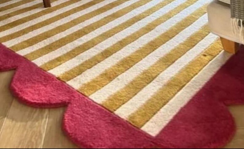 Op de afbeelding: Een gestreept tapijt met afwisselende gouden en witte strepen. Het tapijt heeft een geschulpte, roze rand. Het tapijt ligt op een houten vloer en een deel van een stoelpot is zichtbaar.