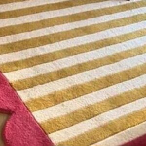 Op de afbeelding: Een close-up van een gestreept tapijt met afwisselend gouden en witte strepen. Het tapijt heeft een geschulpte roze rand. Het tapijt is gemaakt van een zacht materiaal.