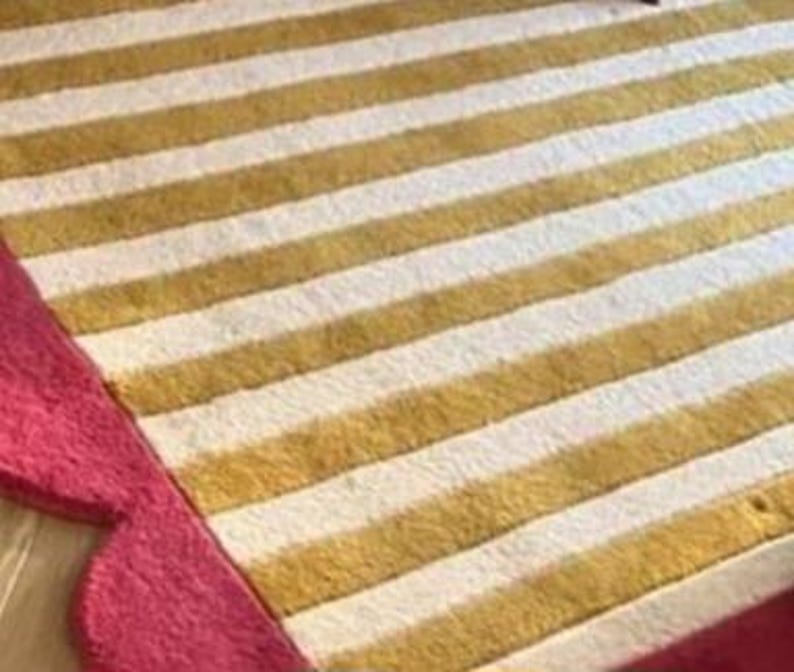 Op de afbeelding: Een close-up van een gestreept tapijt met afwisselend gouden en witte strepen. Het tapijt heeft een geschulpte roze rand. Het tapijt is gemaakt van een zacht materiaal.
